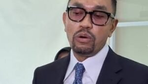 "Potret Ahmad Sahroni mengenakan jas navy, dasi polkadot, dan kacamata hitam, tersenyum lebar saat diwawancarai awak media di lorong Gedung DPR RI pada 19 Februari 2026."