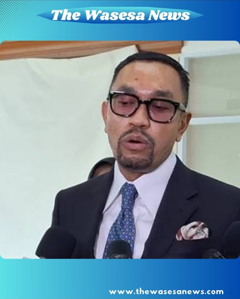 ​"Potret Ahmad Sahroni mengenakan jas navy, dasi polkadot, dan kacamata hitam, tersenyum lebar saat diwawancarai awak media di lorong Gedung DPR RI pada 19 Februari 2026."