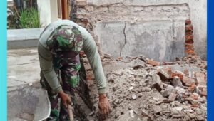 Prajurit TNI sedang menghancurkan sisa-sisa lantai lama menggunakan palu besar dalam kegiatan rehabilitasi rumah milik Bapak Khoirul Anam di Dusun Desa Wonosari, Pasuruan. Di sekitarnya terlihat gerobak dorong dan ember sebagai alat bantu kerja.